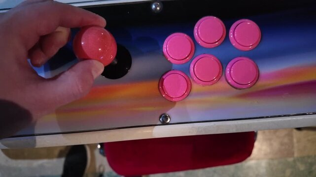 Close-up of a hand holding a red joystick knob next to pink arcade game buttons on a metallic control panel in an indoor setting