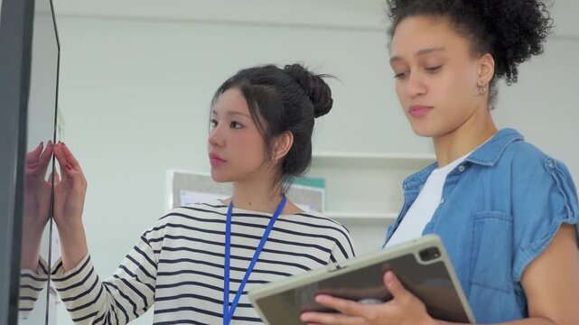 Two focused female colleagues reviewing information together on a tablet, one explaining a point while wearing a lanyard, in a bright collaborative office space.