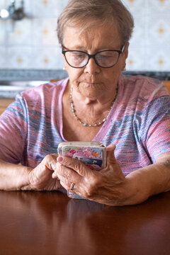 Elderly woman wearing glasses concentrates on her smartphone at a kitchen table, scrolling and texting with a focused expression, showing senior technology use, 