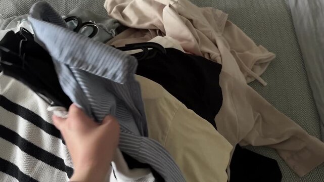 Horizontal close-up video of a hand sorting through black and metal clothes hangers among assorted garments spread across a bed during wardrobe decluttering, closet cleanup and clothing organization