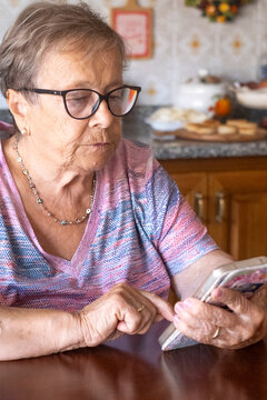 Elderly woman using smartphone at kitchen table, senior grandmother reading messages with glasses and concentrating on touchscreen