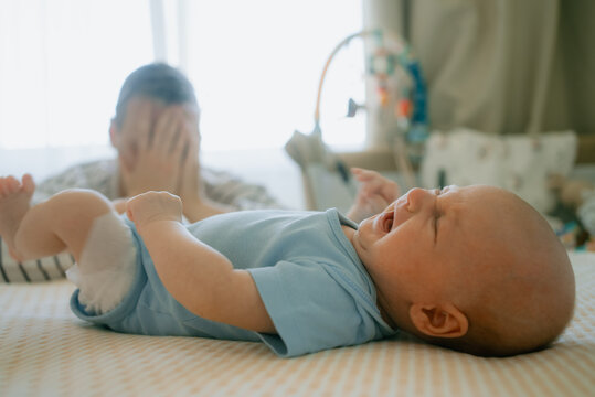 Young father feeling overwhelmed and distressed by a crying infant, covering his face in a gesture of mental exhaustion and postpartum depression while the baby lies upset