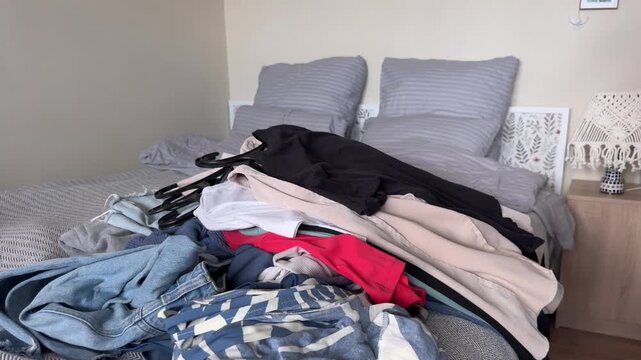 Horizontal video of an unrecognizable person placing another stack of several garments onto a pile of clothes already lying on a bed, adding striped knitwear and dark apparel in a home bedroom