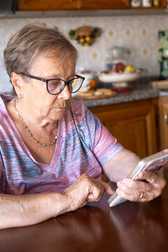 Elderly woman sits at a kitchen table concentrating on her smartphone, tapping the screen with focus. Wearing glasses and a casual patterned shirt, she practices mobile technology