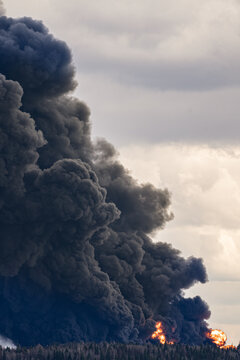 Enormous column of thick black smoke rising from burning car tires over a forest, indicating a large-scale fire at a landfill and generating significant air contamination