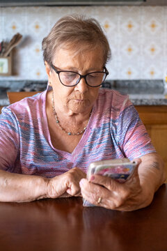 An elderly woman wearing glasses concentrates on her smartphone while sitting at a kitchen table, quietly connecting with family and technology in a domestic, thoughtful moment