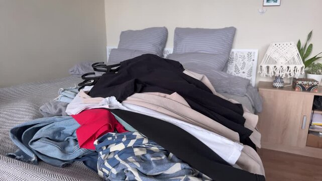 Horizontal video of an unrecognizable person throwing a thick stack of black, beige and white garments onto a pile of assorted clothes spread across a bed, with denim and bright red fabric visible