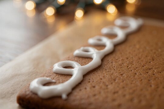 Close-up of decorative white icing swirled on a gingerbread wall panel, suggesting the making of a gingerbread house with warm bokeh lights in the background.