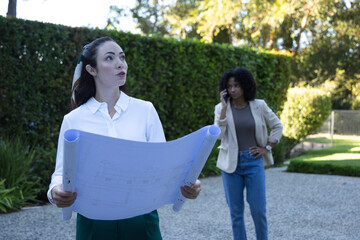 Naklejka premium Asian woman in blouse holding plans on gravel drive, assessing site while colleague on smartphone
