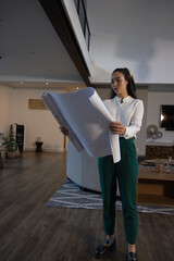 Fototapeta premium Adult female holding blueprints, studying modern living room, wearing white collared blouse