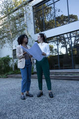 Diverse female coworkers reviewing large blueprints in courtyard by reflective glass windows © WavebreakMediaMicro