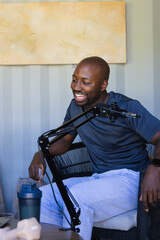 African man smiling, speaking, leaning in woven armchair with microphone boom arm and tumbler © WavebreakMediaMicro
