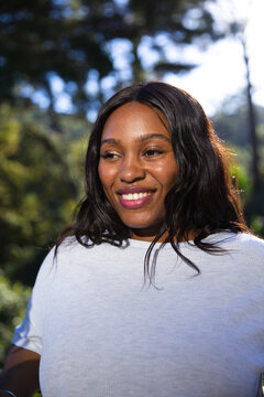 African adult woman smiling while standing in woodland wearing white t-shirt, showing sunlit bokeh