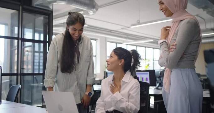 Diverse female team watching seated colleague gesturing at laptop, reviewing data, noting scarf