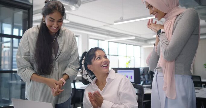 Diverse female team celebrating win, seated woman pointing at laptop and highfiving in open office