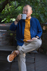 Senior Asian man sitting on wooden bench on deck sipping from grey ceramic mug wearing wristwatch © WavebreakMediaMicro