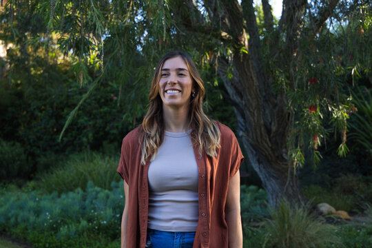 Adult woman standing in lush garden wearing rust shirt and jeans by large tree and rocks