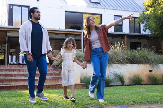 Diverse family with child walking across front lawn of modern house, holding hands, mother pointing