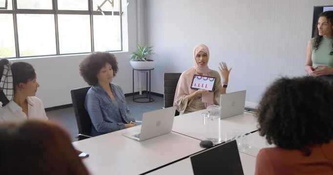 Female presenter wearing light scarf raising tablet showing charts in conference room seeking input