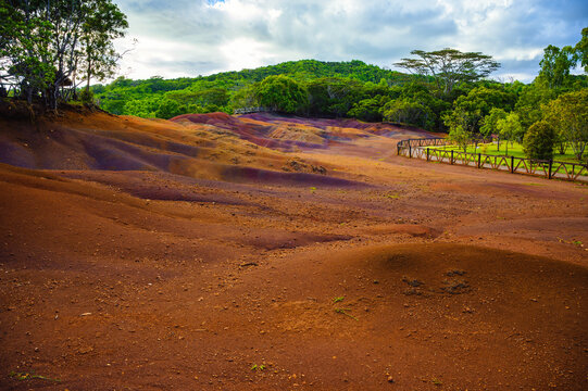 Vibrant red, brown, and purple sand dunes at Seven Colored Earth in Chamarel Geopark, Mauritius, bordered by tropical rainforest under dramatic clouds.
