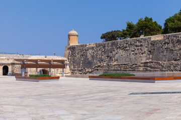 Historic stone fortress with a rounded watchtower overlooking a wide sunlit plaza and coastal ramparts, framed by clear blue sky and scattered trees. Valletta, Malta © evannovostro