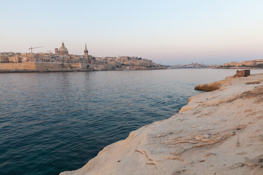 Golden evening light illuminates Valletta historic limestone skyline and domed churches across the calm Mediterranean harbor, framed by rocky sandstone foreground and a tranquil pastel sky at sunset