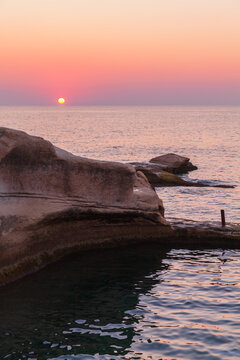 Soft pink and orange sunset over a rocky coastline with calm sea and reflective water at dusk, evoking peaceful, tranquil feelings and coastal serenity ideal for travel, nature and relaxation themes