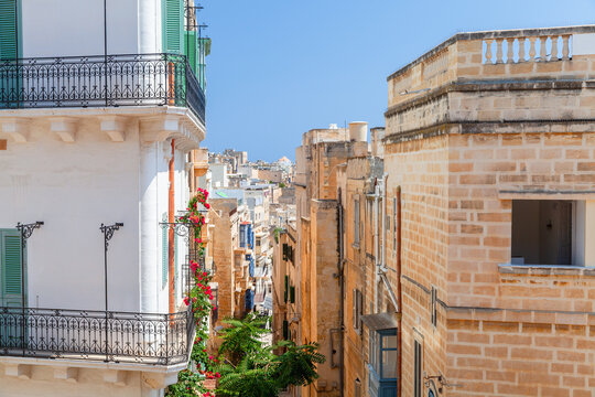 Sunlit Mediterranean scene of balconies with green shutters and wrought-iron railings overlooking a narrow historic limestone street of Valletta, Malta