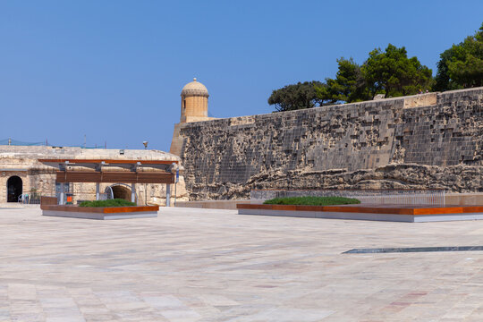 Historic stone fortress with a rounded watchtower overlooking a wide sunlit plaza and coastal ramparts, framed by clear blue sky and scattered trees. Valletta, Malta