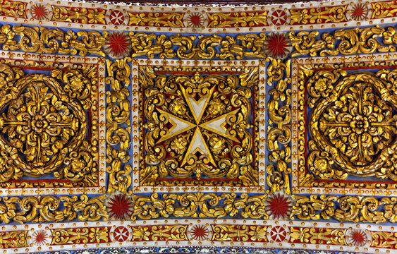 Saint John Co-Cathedral decoration, Valletta, Malta. Ornate gilded ceiling panel featuring a central Maltese cross, intricate gold leaf scrollwork and floral motifs in red and blue accents