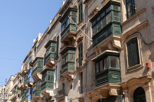 Rows of traditional Maltese wooden balconies and shuttered windows line a historic Valletta street, showcasing warm sandstone facades, architectural detail and Mediterranean charm under clear blue sky