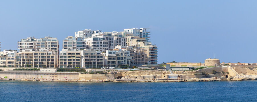 A cluster of modern waterfront apartment buildings sits behind a low historic stone promontory, overlooking a calm blue sea and rocky shoreline under a clear sky. Sliema, Malta