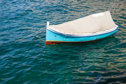 Small blue rowboat with canvas cover moored on calm teal water, casting sunlit reflections and gentle ripples. Nautical, serene scene evoking summer leisure, solitude, travel and coastal charm, Malta