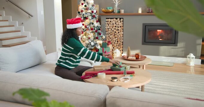 African-American woman at home wearing red hat wrapping Christmas gifts on table with red paper