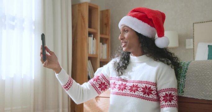 African American woman in Santa hat tapping smartphone starting call on bed sharing wrapped gift