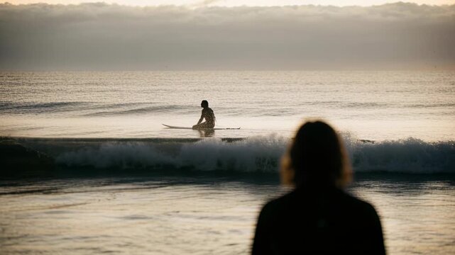 Watching observer in wetsuit at shore, seated surfer shifting on longboard, paddling to ride swell