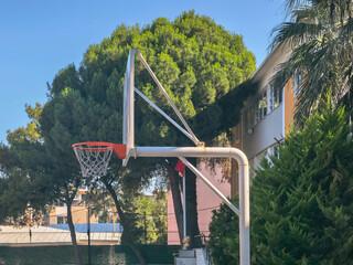 A white metal basketball hoop with a backboard and net on an outdoor asphalt school sports court, surrounded by green trees. © Mariia135
