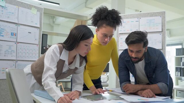 Three colleagues huddled closely together, reviewing a data analytics chart on a tablet screen, deeply focused and collaborative moment in a busy modern office.