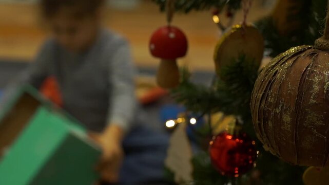 Close-up shot of Christmas tree ornaments and lights in focus, while a blurred toddler opens and unwraps gift boxes in the background living room.