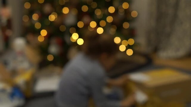 Out-of-focus background shot showing beautiful warm Christmas tree bokeh lights with a blurred toddler sitting on the carpet opening present boxes.