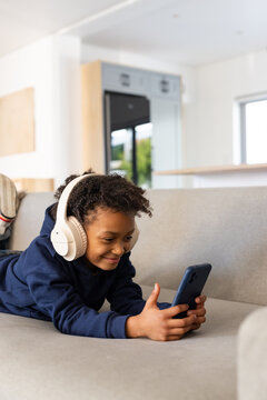 Female child lying on grey sofa at home wearing navy hoodie cream headphones holding smartphone