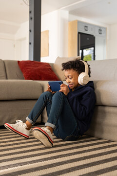 African American girl sitting on rug in navy hoodie with headphones holding blue smartphone smiling