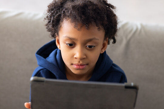African American female child sitting on light-gray sofa at home wearing navy hoodie holding tablet