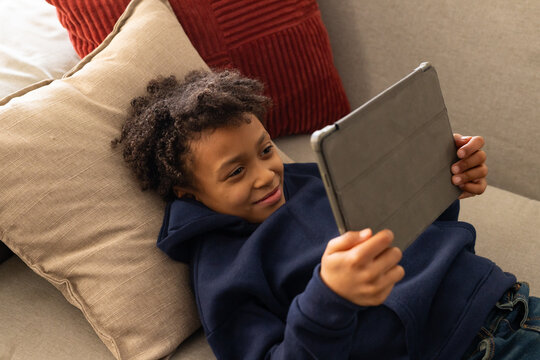 African American youth female reclining on beige sofa holding gray tablet, in navy hoodie and jeans