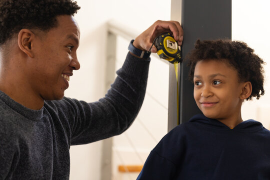African American father measuring son against post in hallway near staircase, holding tape measure