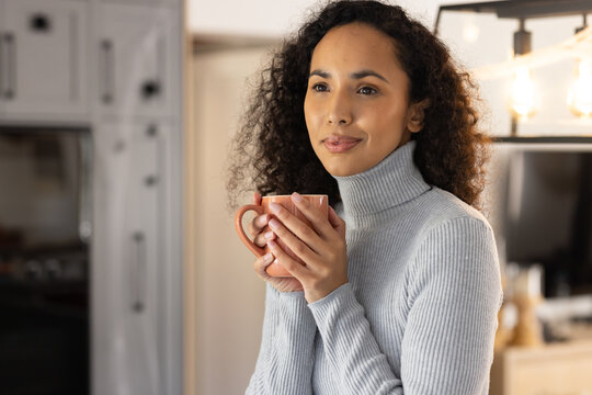 African American woman holding ceramic mug near chest, wearing gray ribbed sweater in home kitchen