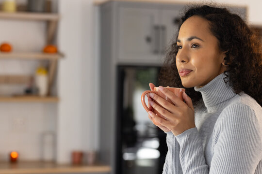 African American woman posing in kitchen holding peach mug by face wearing gray sweater, copy space