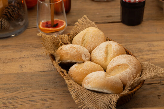 Woven basket is holding six floured rolls resting on worn wood table with citrus, pinecones, jam