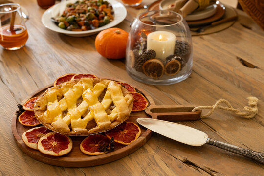 Lattice-top fruit pie is resting on round wooden board on rustic dining table with dried citrus