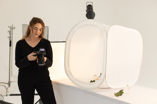 Woman photographer in black checking DSLR near light tent bottles on studio tabletop, copy space